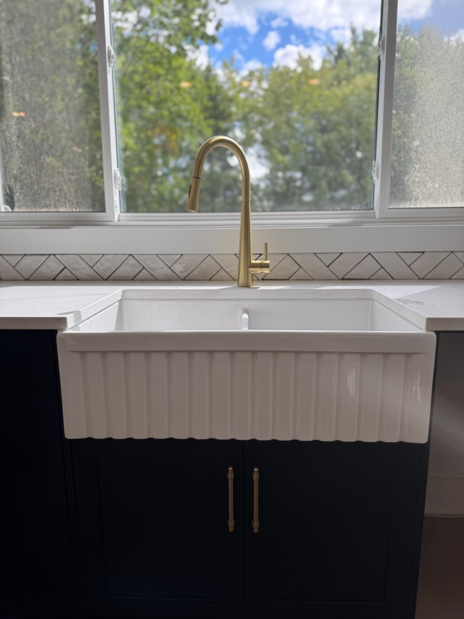 Fluted farmhouse sink with a brass faucet under a window onto trees
